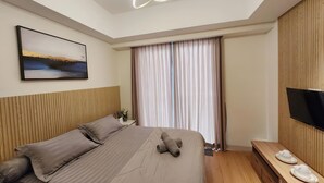 Une chambre d’hôtel moderne avec un grand lit, un téléviseur à écran plat, une tête de lit en bois, un tableau au mur et une fenêtre avec des rideaux.
