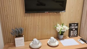 Un bureau en bois avec un téléviseur à écran plat fixé au-dessus. Sur le bureau, on trouve une petite plante en pot, deux chapeaux en papier blancs, un bloc-notes blanc, un stylo et une affiche motivante.