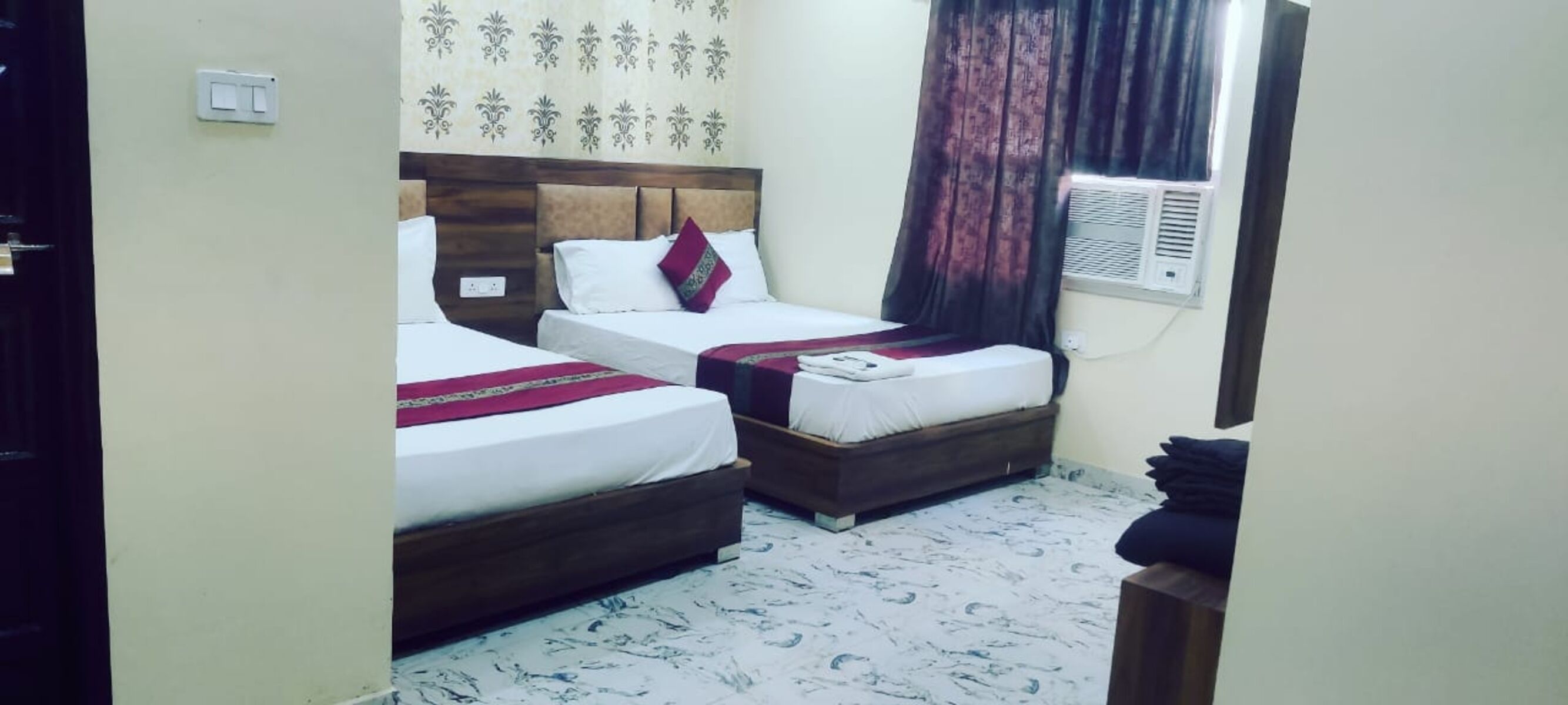 A hotel room with two beds, a wooden headboard, a patterned curtain, and a wall-mounted air conditioning unit.