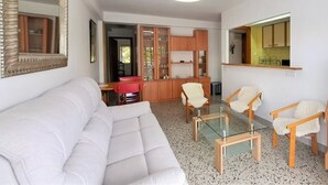 A modern living room with a white sofa, glass coffee table, and wooden chairs. There is a kitchen area with a sink and cabinets. - Family apartment in Salou with Wi-Fi 300m from the beach (Salou)