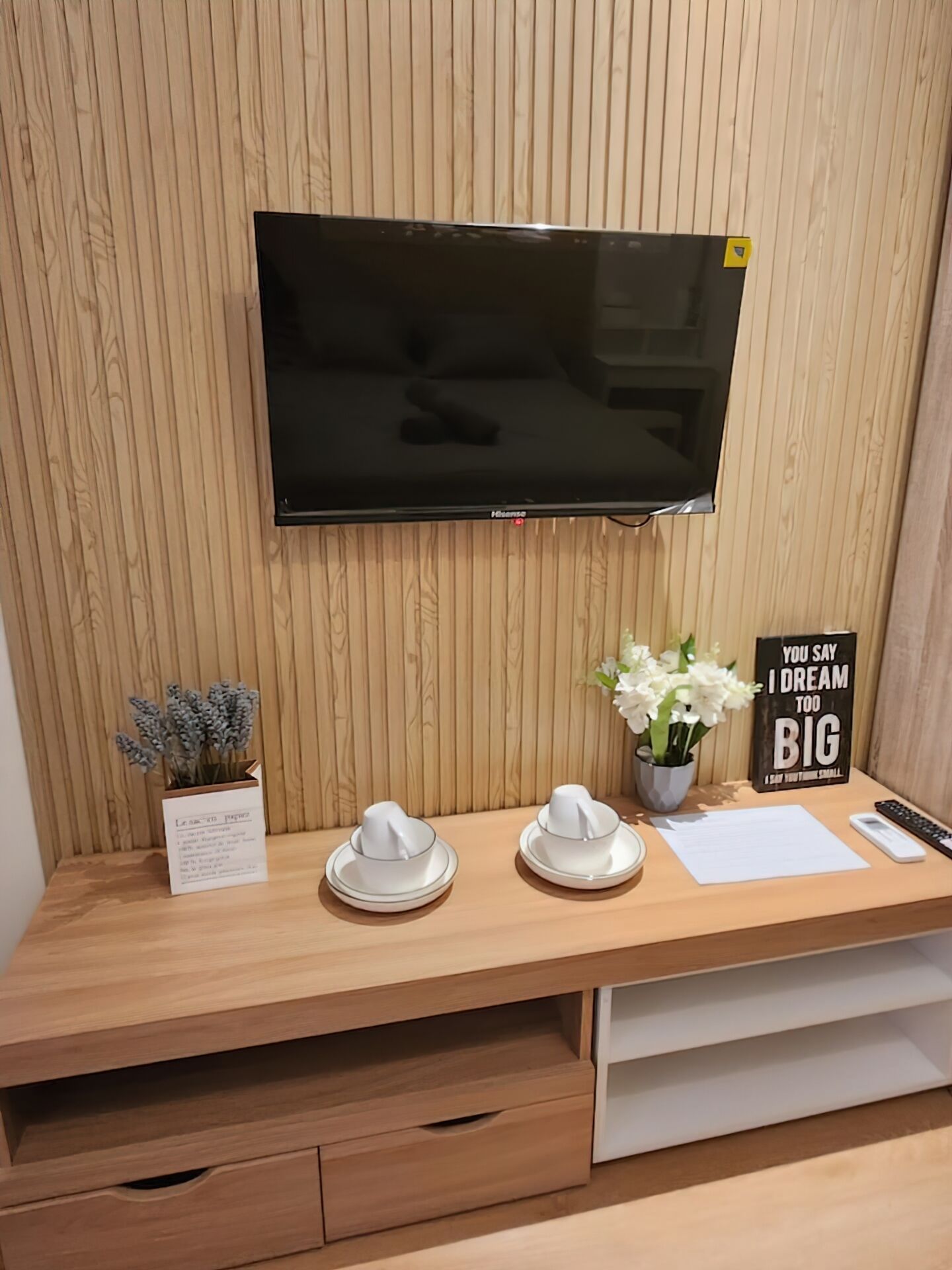 Un escritorio de madera con un televisor de pantalla plana montado encima. Sobre el escritorio, hay una pequeña planta en maceta, dos gorros de papel blancos, un bloc de notas blanco, un bolígrafo y un cartel motivacional.