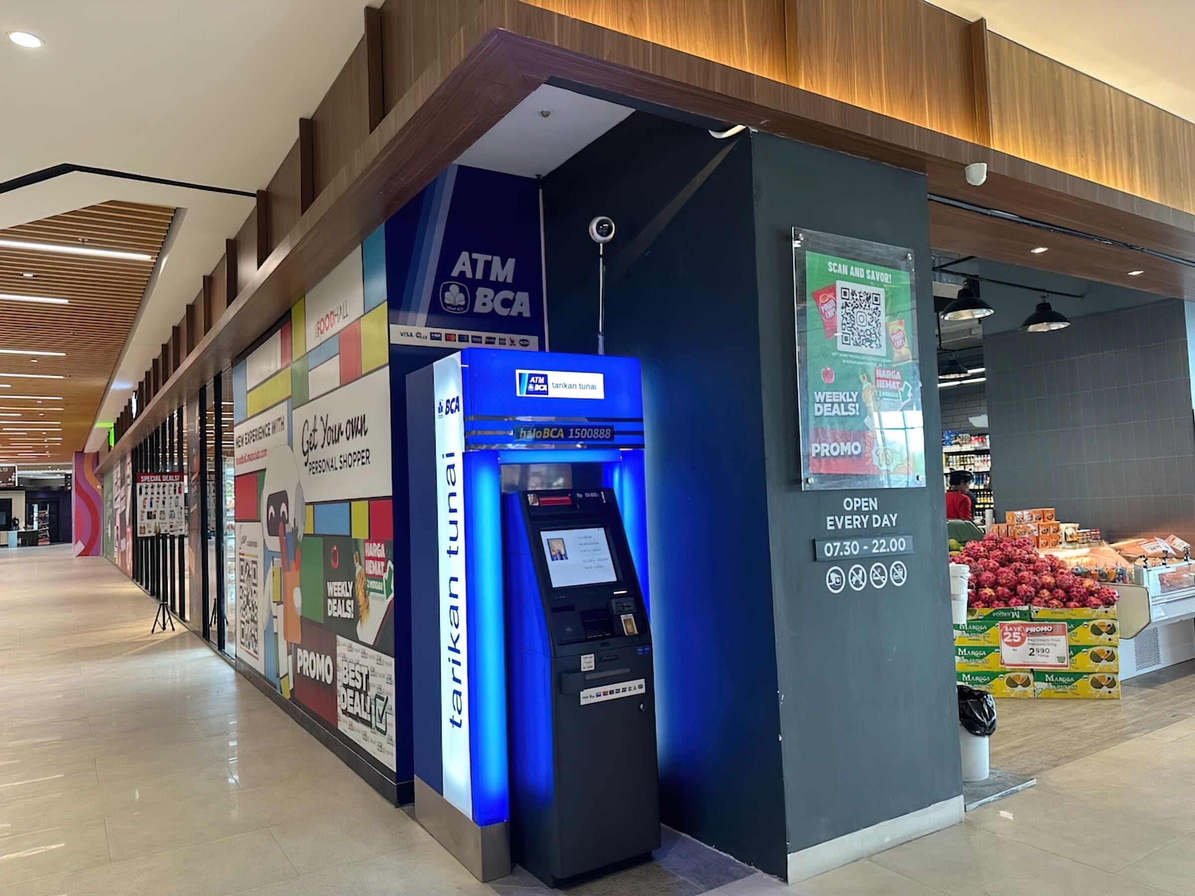 Un distributeur automatique de billets arborant le logo de BCA, une affiche promotionnelle pour les offres hebdomadaires et un présentoir de fruits dans une zone commerciale.