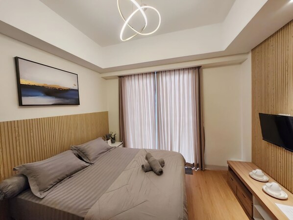 Une chambre d’hôtel moderne avec un grand lit, un téléviseur à écran plat, une tête de lit en bois, un tableau au mur et une fenêtre avec des rideaux.