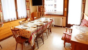 Una zona de menjador amb taula preparada per a sis, cadires de fusta, un televisor a la paret i grans finestres amb cortines.