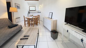 Un salon avec un canapé, une télévision et des télécommandes, un coin repas avec des chaises et une table, et une cuisine à l’arrière-plan.