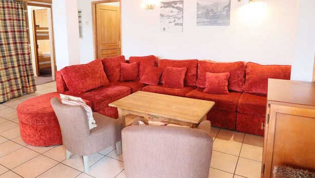 Un salon chaleureux avec un canapé d’angle rouge, une table basse en bois et deux fauteuils gris.