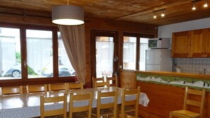 Un intérieur en bois comprenant une table et des chaises, un coin cuisine avec des placards, et une vue sur l’extérieur grâce à de grandes fenêtres.