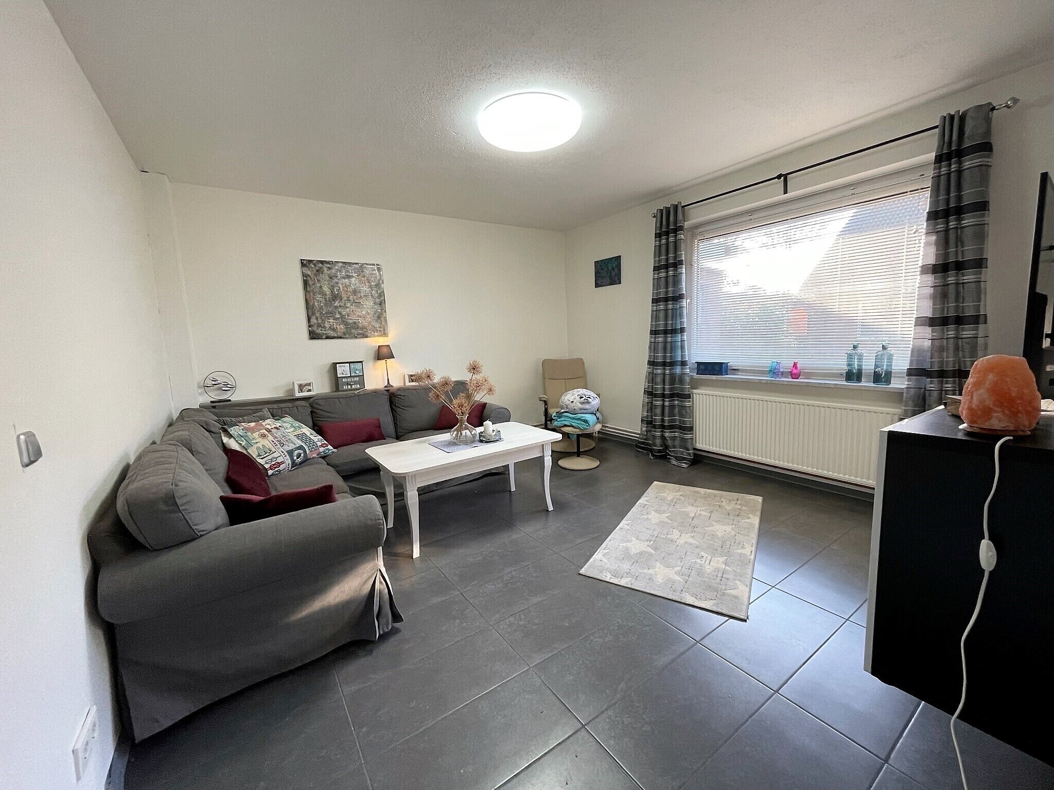 Ein Wohnzimmer mit grauem Sofa, einem weißen Couchtisch, einem kleinen Beistelltisch, einer Heizung und einem Fenster mit Vorhängen.