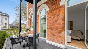 A modern hotel room with a large window, a wooden deck, and a view of a historic building. - Luxury apartment with terrace, free SPA use and gym (Sylt)