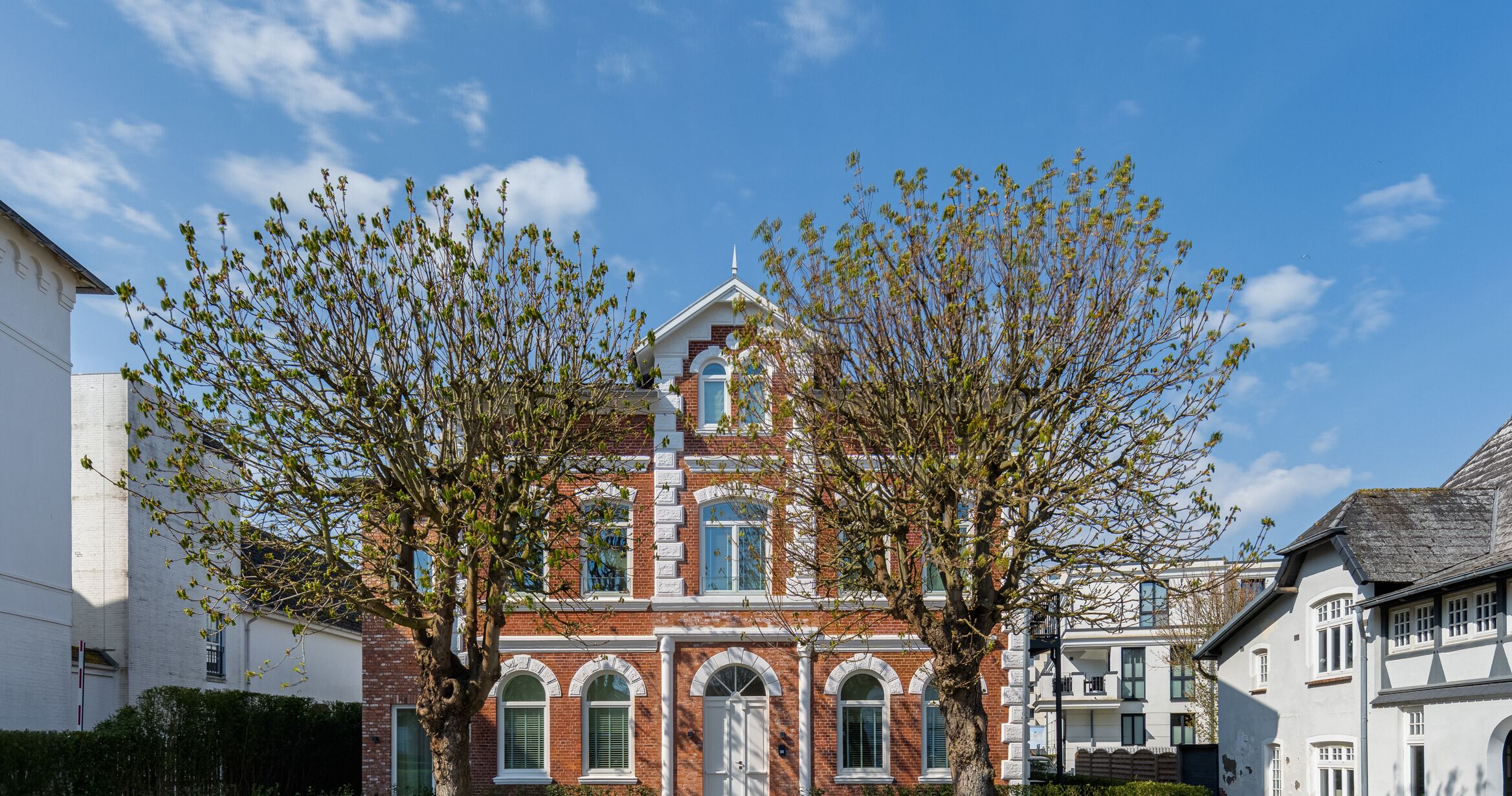 A traditional brick building with a central entrance, flanked by two trees with budding leaves.
