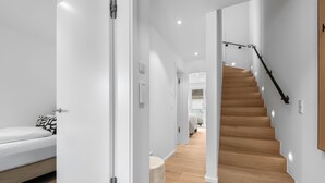 Un intérieur moderne avec un escalier en bois, un lit agrémenté d’oreillers à motifs et un couloir menant à une autre pièce.