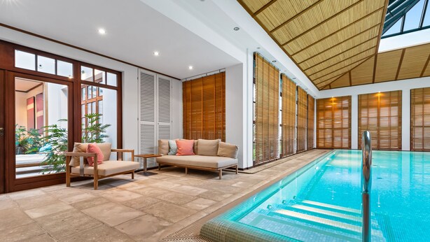 Un espace piscine intérieur moderne, doté d’un canapé et de fauteuils, de stores en bois et d’une porte vitrée donnant sur l’extérieur.