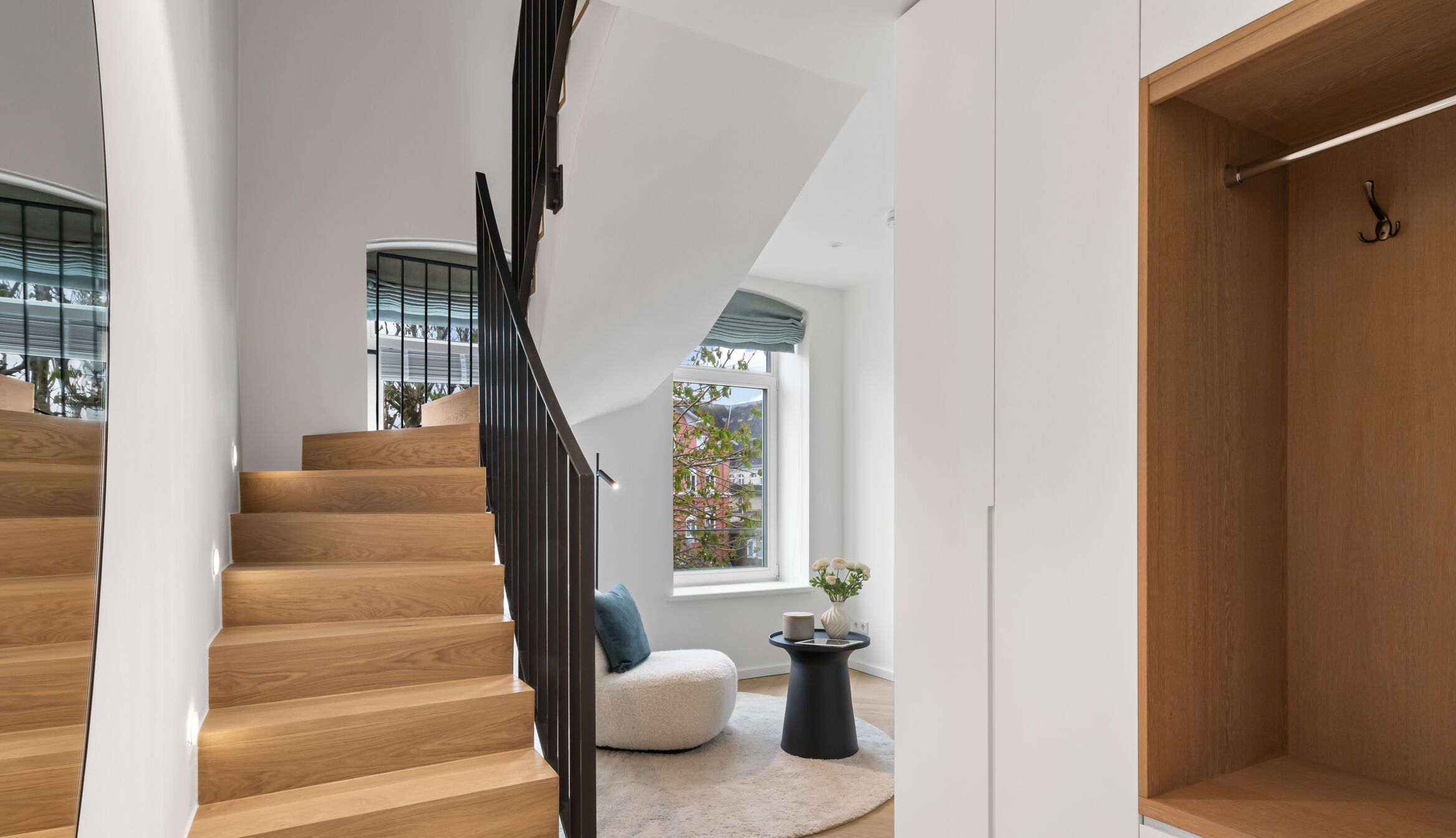 Una escalera moderna con escalones de madera y barandillas negras que conduce a una habitación con pared blanca, una ventana con vistas a edificios y una pequeña mesa redonda con un florero y una planta.
