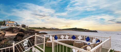 A seaside terrace with a circular seating area, a staircase leading down to the beach, and a view of the ocean and sunset.