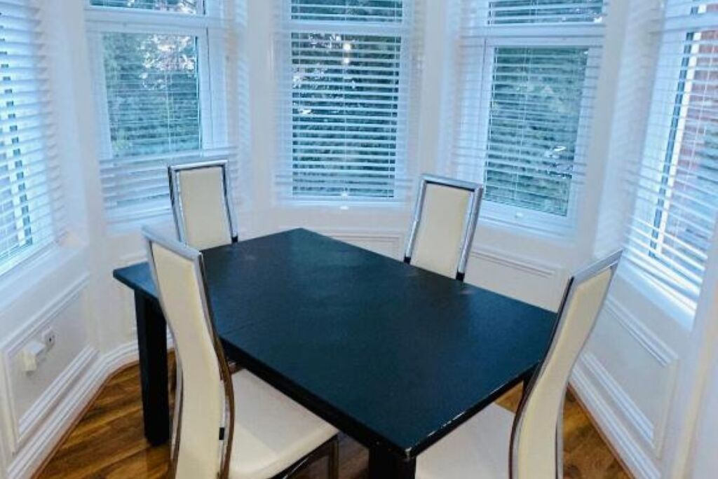 Un coin repas avec une table noire et des chaises blanches, entouré de grandes fenêtres équipées de stores.