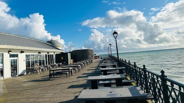 Une jetée en bois, dotée de bancs et d’une rambarde, mène à un bâtiment dont le toit est en pente et qui possède de grandes fenêtres.