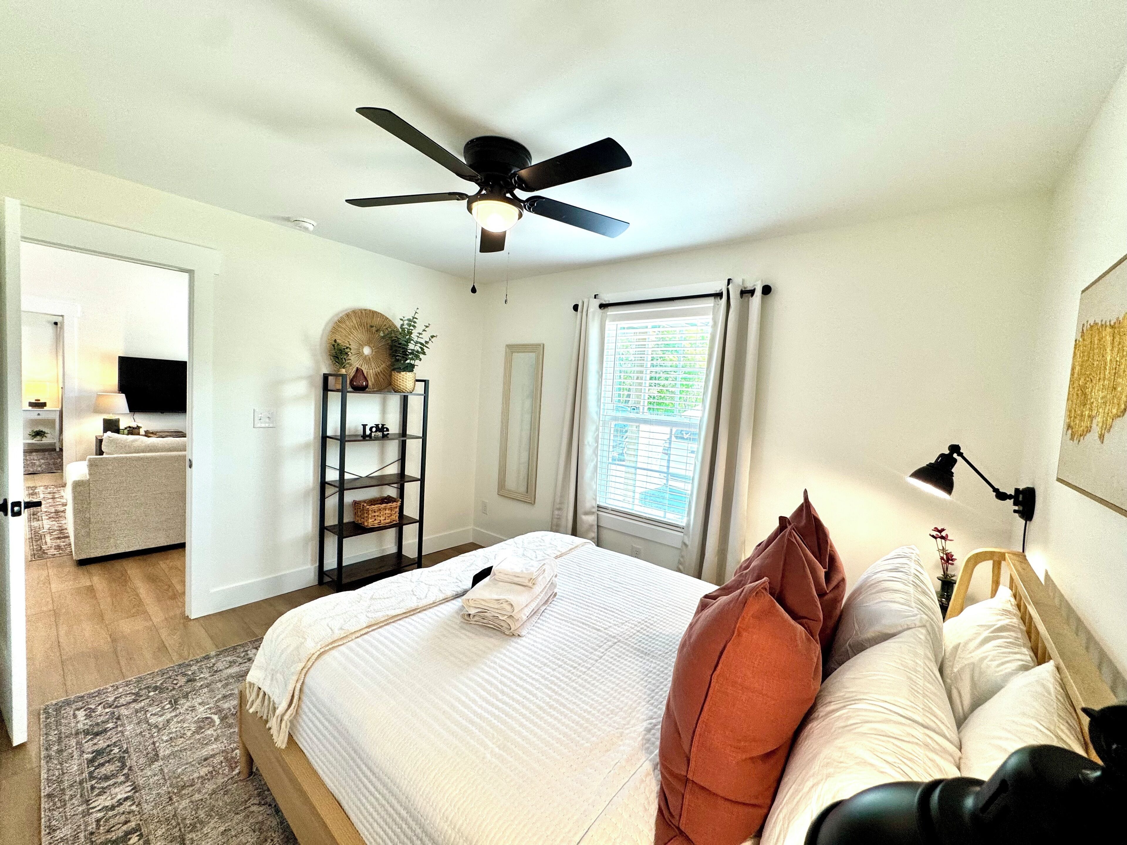 A bedroom with a bed, a ceiling fan, a window with curtains, a bedside table with a lamp, and a shelf with decorative items.