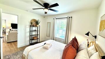 A bedroom with a bed, a ceiling fan, a window with curtains, a bedside table with a lamp, and a shelf with decorative items.