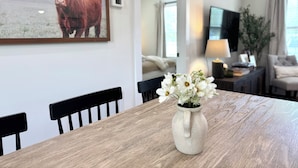 A dining area with a wooden table, chairs, a vase with flowers, a framed picture of a bull, and a living room in the background.