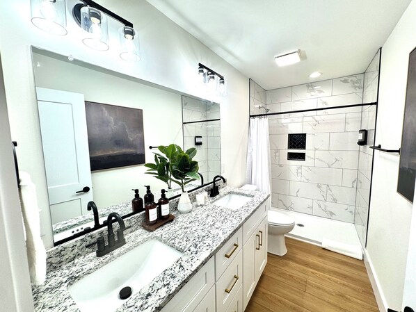 Un baño moderno con encimera de granito, un espejo grande, ducha con puerta de cristal y un inodoro.