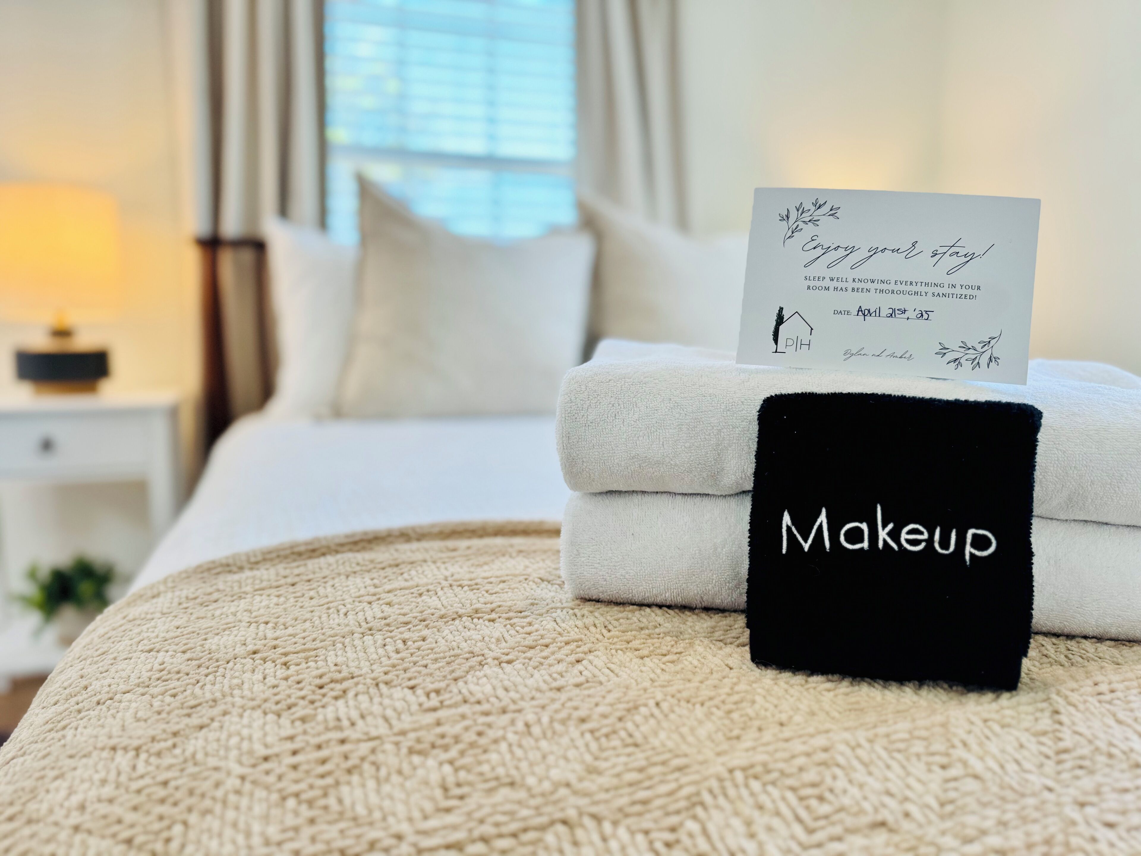 A neatly made bed with folded towels and a black towel with "Makeup" written on it, a card on the bed, and a nightstand with a lamp in a hotel room.