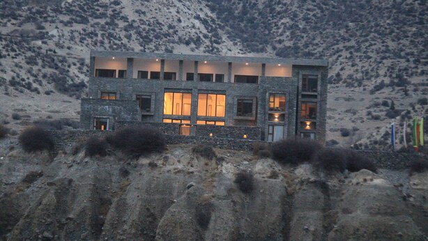 Un edifici modern amb nombroses finestres i façana de pedra, davant d'un paisatge de muntanyes.
