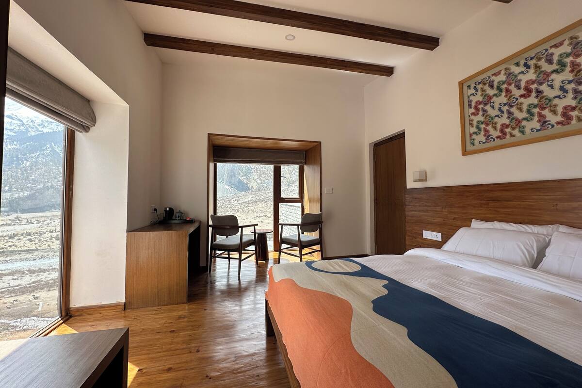 A bedroom with a large bed, wooden beams on the ceiling, a desk with a chair, and a view of a mountainous landscape through a window.