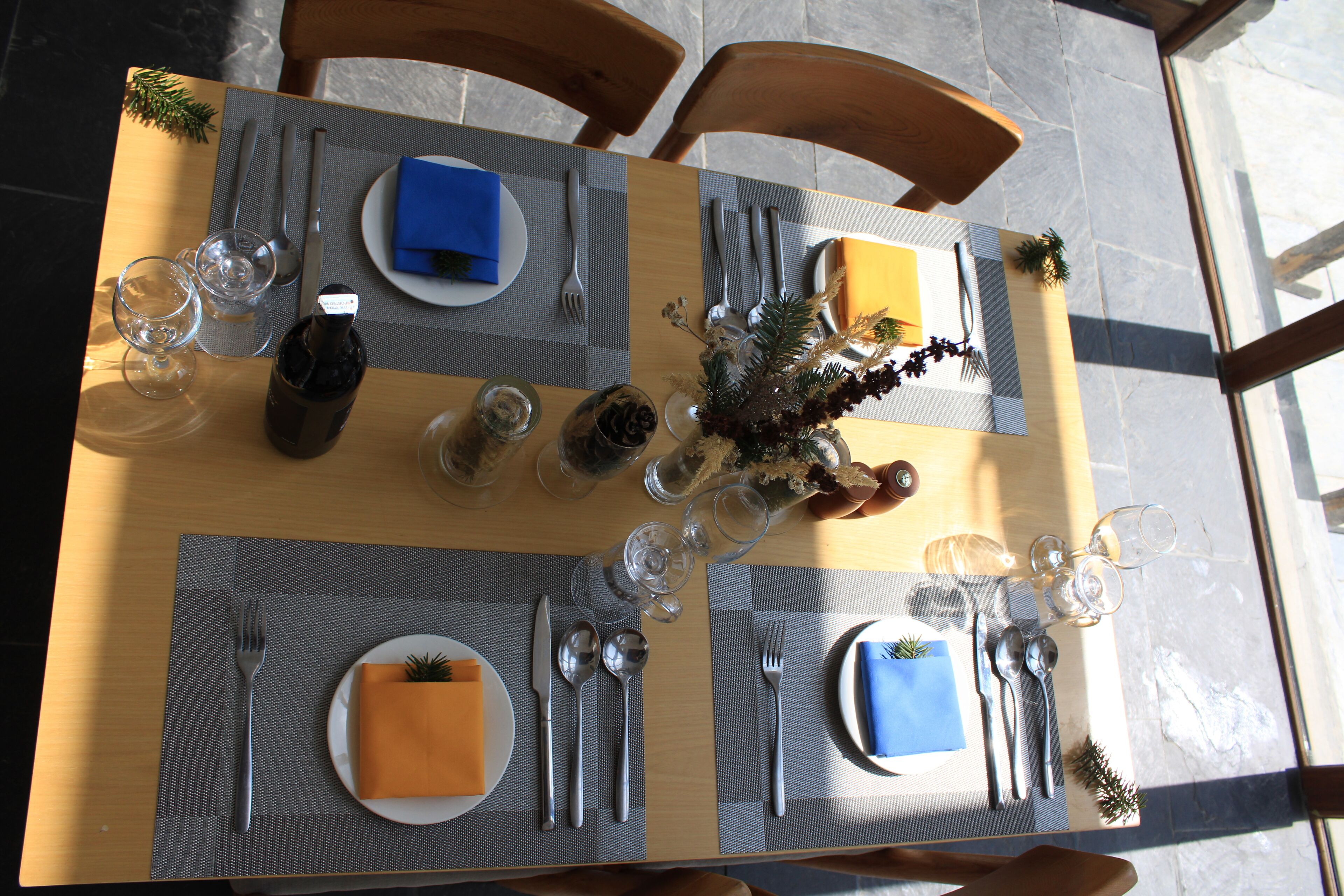 A dining table set with plates, cutlery, glasses, and a bottle, with a vase of greenery in the center.