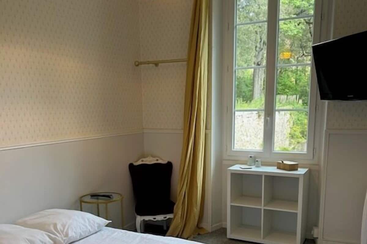 A neatly made bed with pillows and a folded towel, a bedside table with a lamp, a window with curtains, and a TV mounted on the wall.