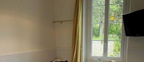 A neatly made bed with pillows and a folded towel, a bedside table with a lamp, a window with curtains, and a TV mounted on the wall.