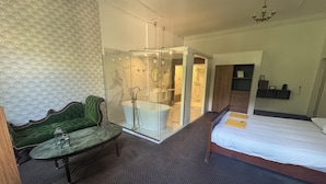 Une chambre d’hôtel avec un lit, un fauteuil recouvert d’un tissu vert, une douche séparée par des parois en verre et un mur décoré d’un motif imitant le marbre.
