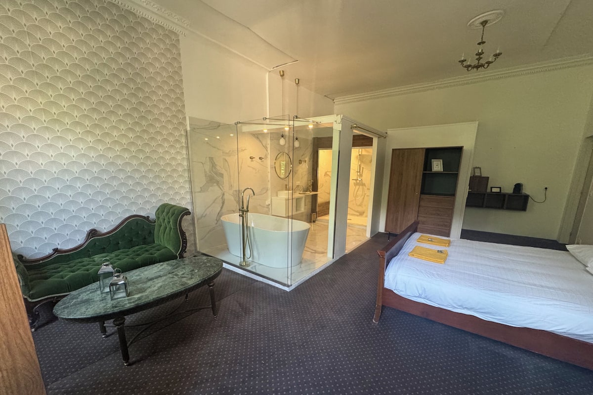A hotel room with a bed, a green upholstered chair, a glass-enclosed shower area, and a marble-patterned wall.
