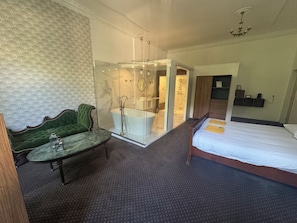 Une chambre d’hôtel avec un lit, un fauteuil recouvert d’un tissu vert, une douche séparée par des parois en verre et un mur décoré d’un motif imitant le marbre.