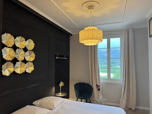Une chambre d’hôtel avec un lit, un élément décoratif mural, un fauteuil vert, une petite table avec un vase et une vue sur les montagnes par la fenêtre.