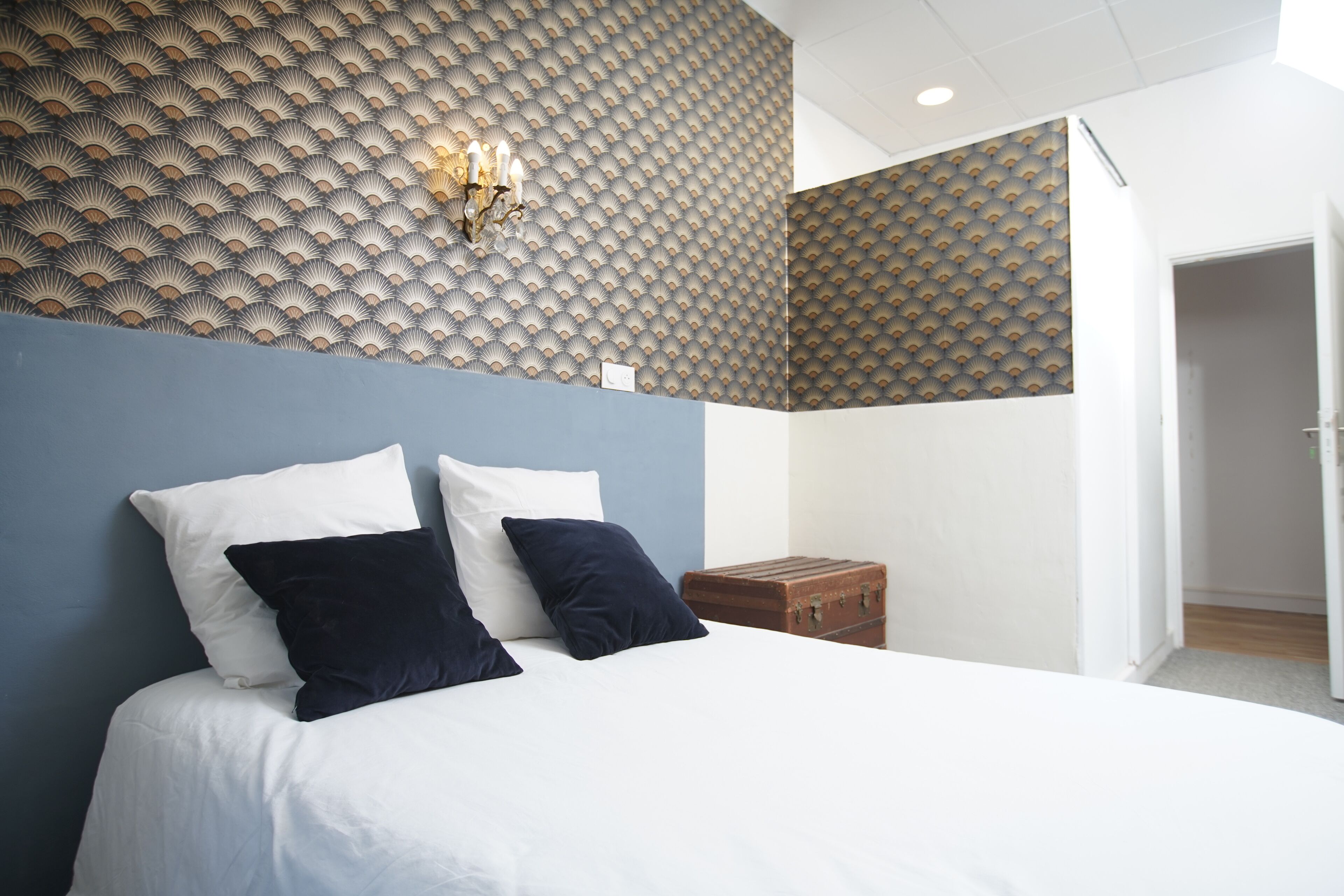 Een net opgemaakt bed met witte en marineblauwe kussens, een behang met patroon en een klein houten dressoir.