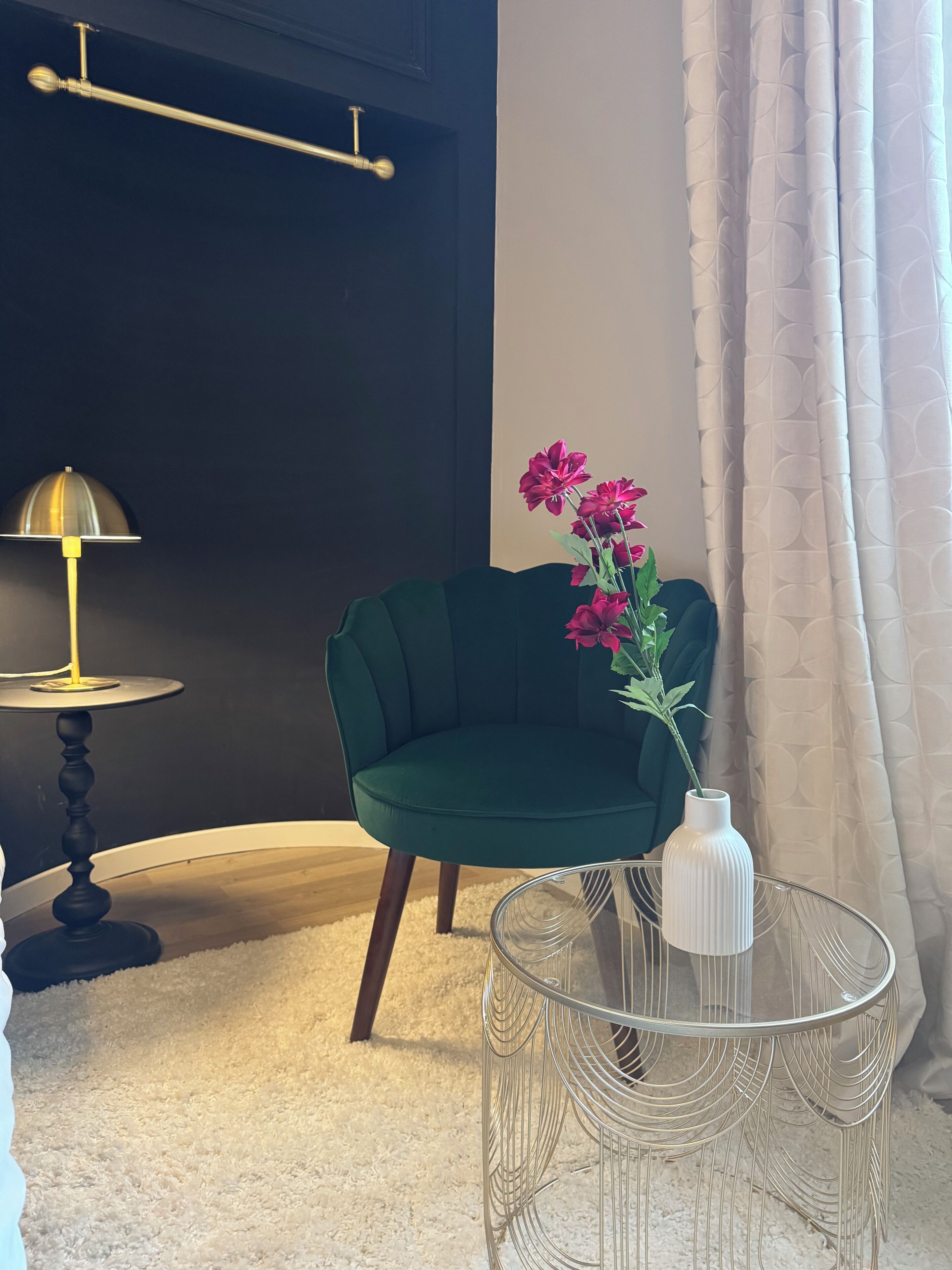 A room with a dark wall, a green chair, a white vase with flowers, a round glass table, and a floor lamp.