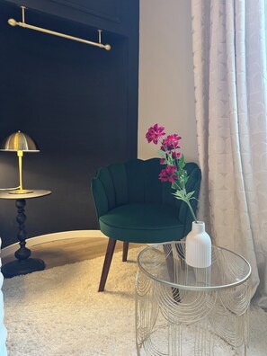 Une pièce avec un mur de couleur sombre, un fauteuil vert, un vase blanc rempli de fleurs, une table ronde en verre et un lampadaire.