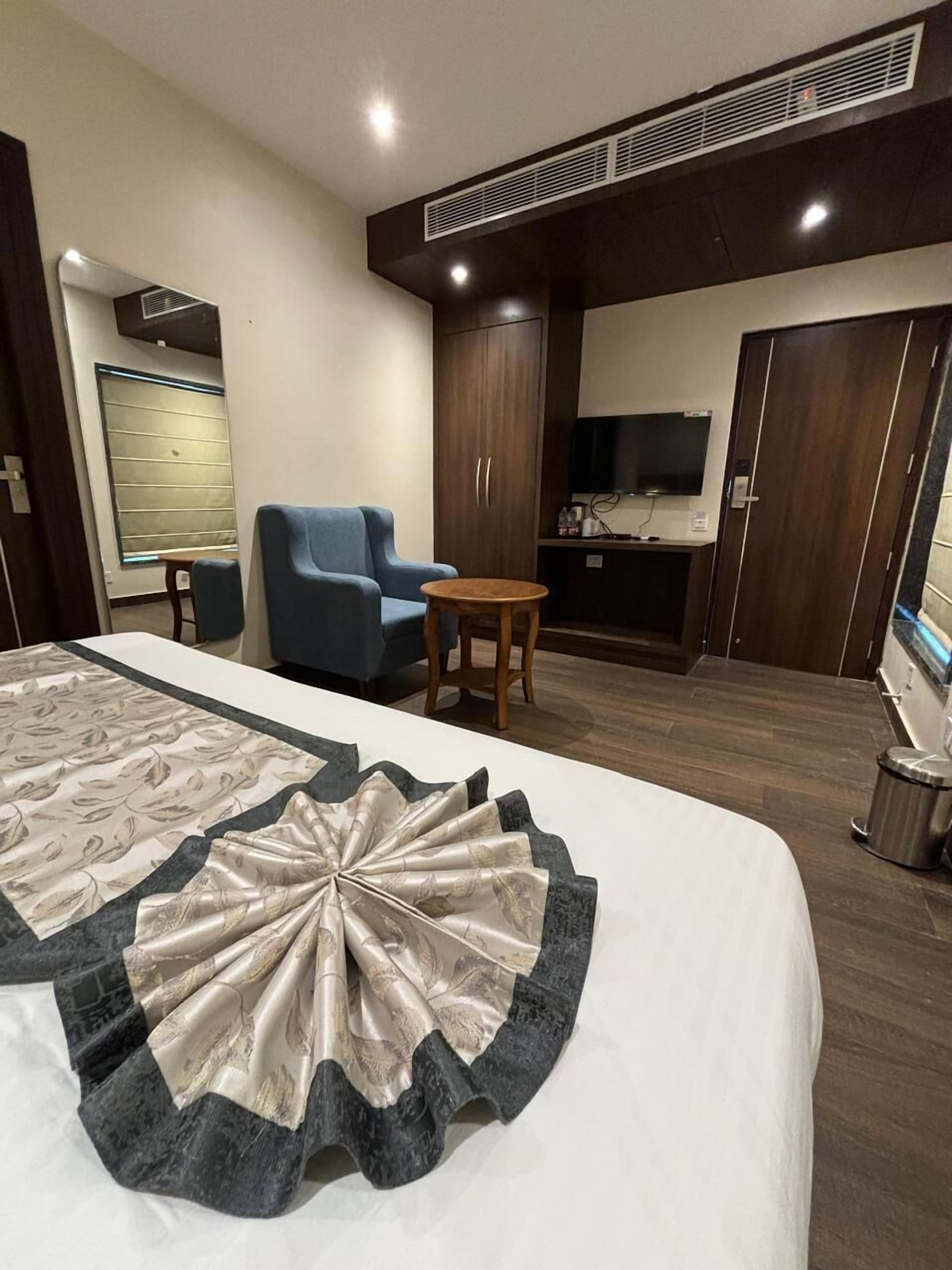 Una habitación de hotel con una cama grande, un sillón azul, una pequeña mesa de madera, un televisor y una zona de kitchenette.
