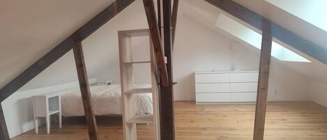 A high-ceilinged attic bedroom with exposed wooden beams, a white bed, a white dresser, and a small white nightstand.