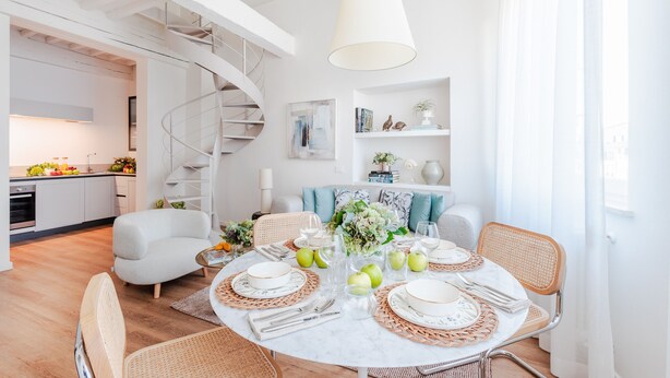 A modern dining area with a round table set for four, surrounded by wicker chairs. A spiral staircase is visible in the background, leading to an upper level. - Letizia Penthouse in Lucca (Lucca)