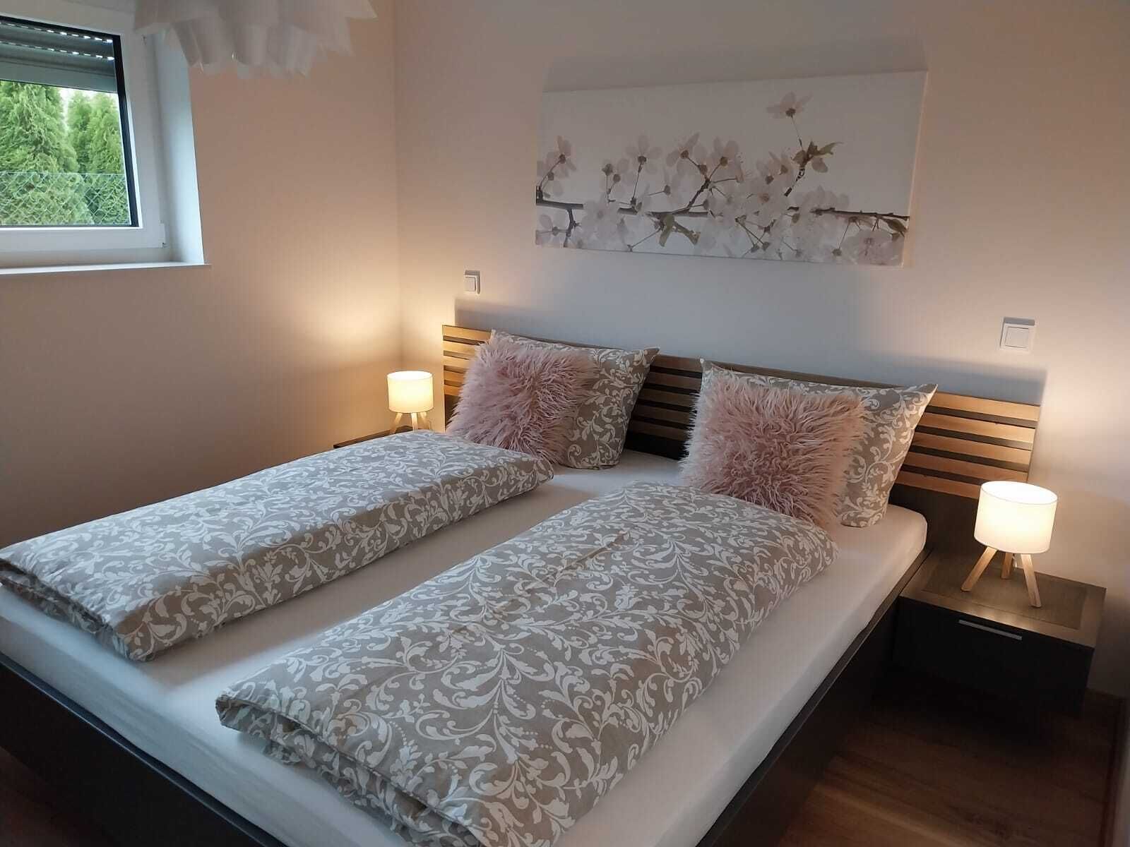 A double bed with patterned bedding, two nightstands with lamps, a window with blinds, and a wall-mounted floral artwork.