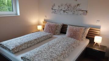 A double bed with patterned bedding, two nightstands with lamps, a window with blinds, and a wall-mounted floral artwork.