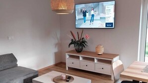 Una sala moderna con un sofá gris, una mesa de centro blanca y una mesa de comedor de madera. Un televisor está montado en la pared, y hay una planta sobre la mesa de centro.