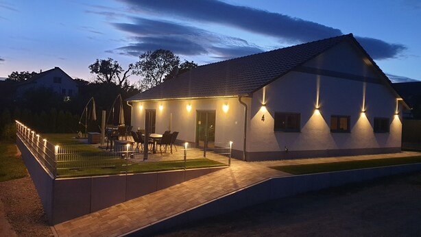 Una casa moderna con porche cubierto, mobiliario exterior y fachada iluminada al anochecer.