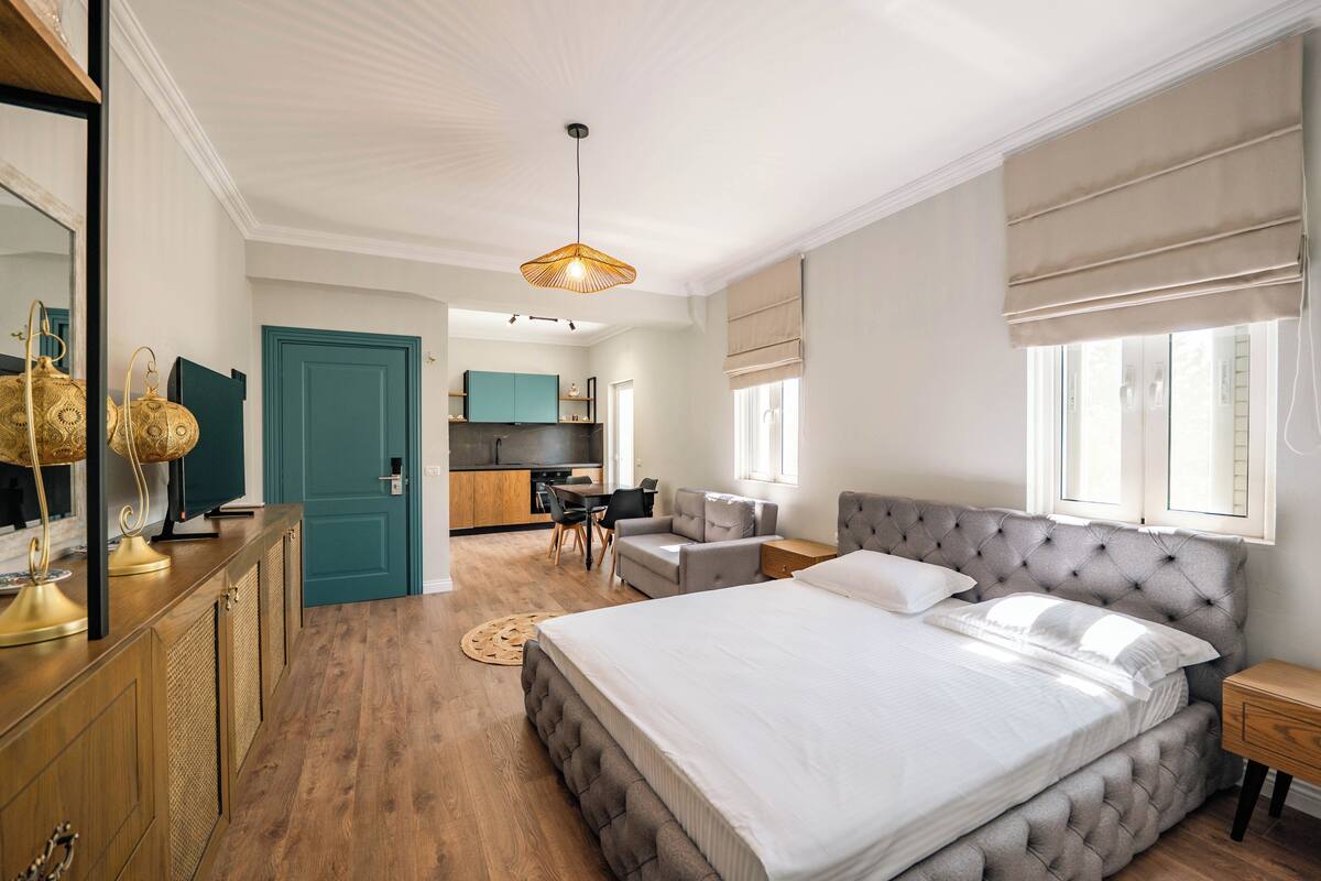 A modern bedroom with a large bed, a desk, and a kitchenette in the background.