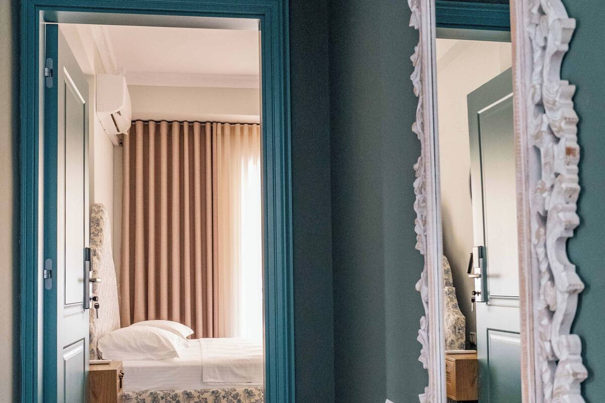 A hallway with a bed in the background, a mirror with an ornate frame, and a woven rug on the floor.