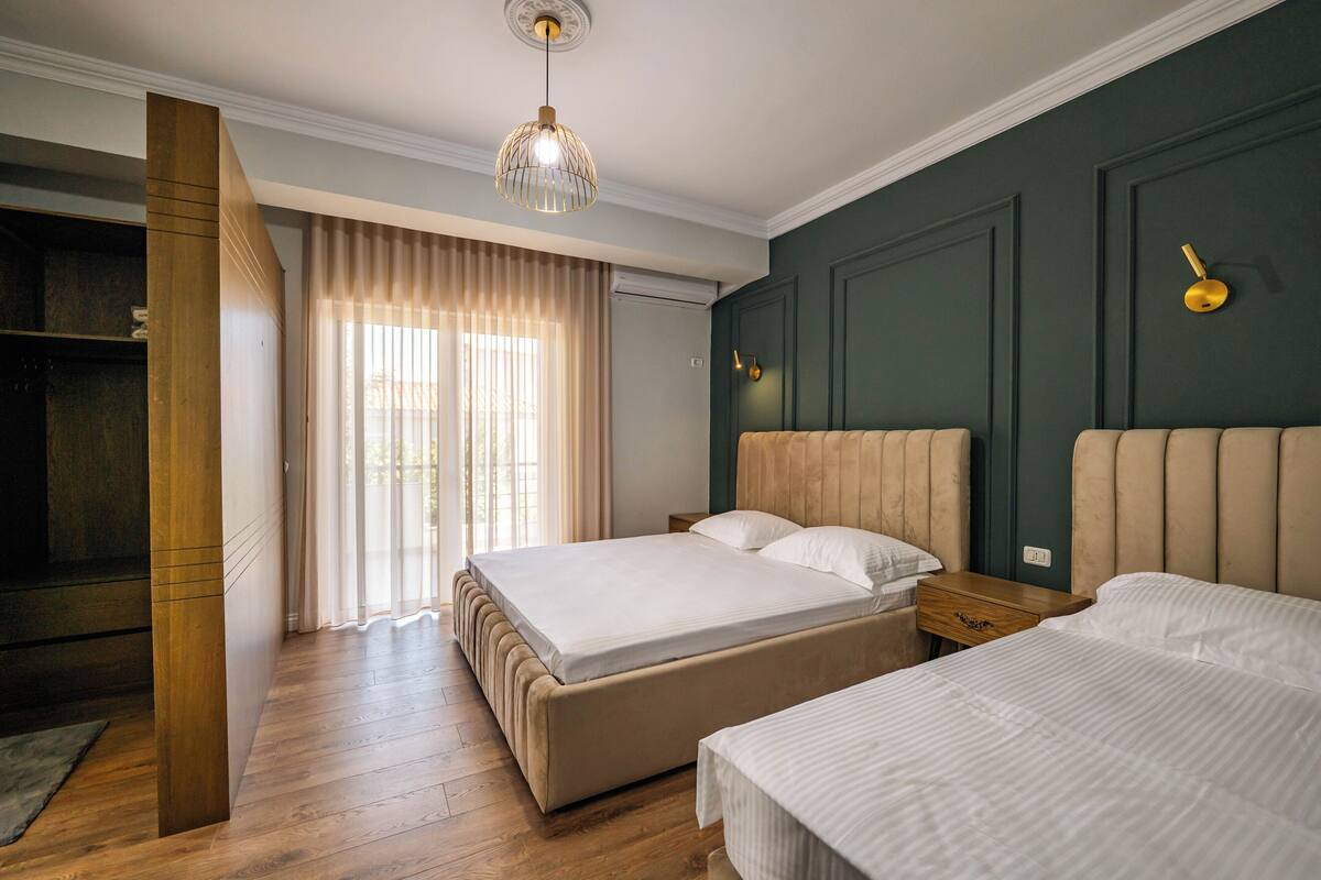A bedroom with two beds, a wooden floor, a large window with curtains, a hanging light fixture, and a wall clock.