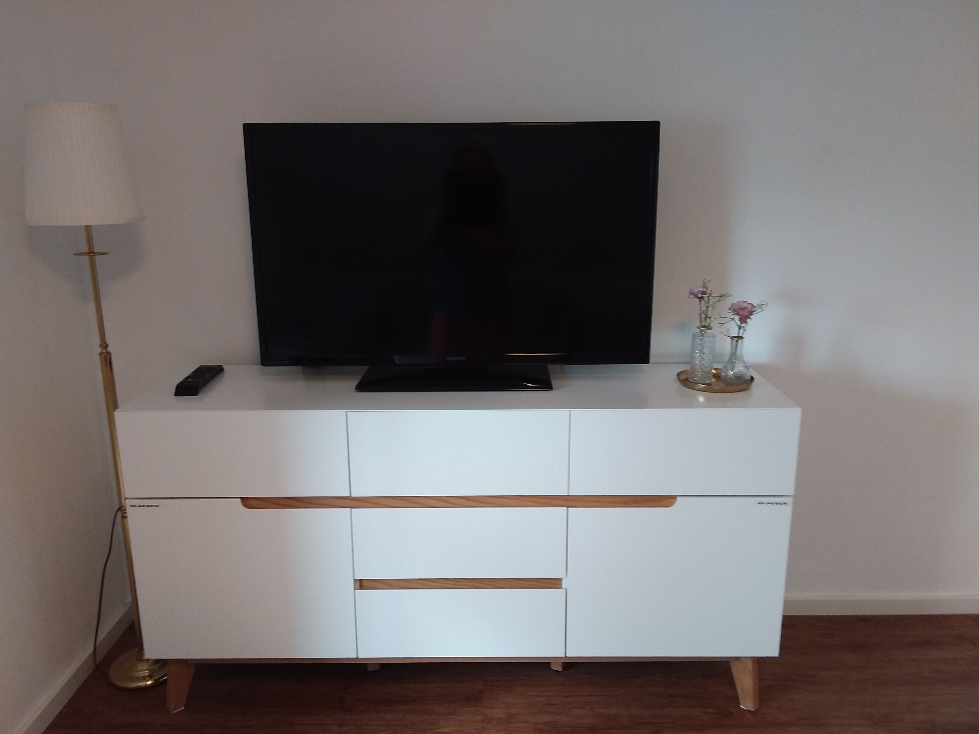 A white entertainment center with a flat-screen TV, a remote control, and a vase with flowers on top.
