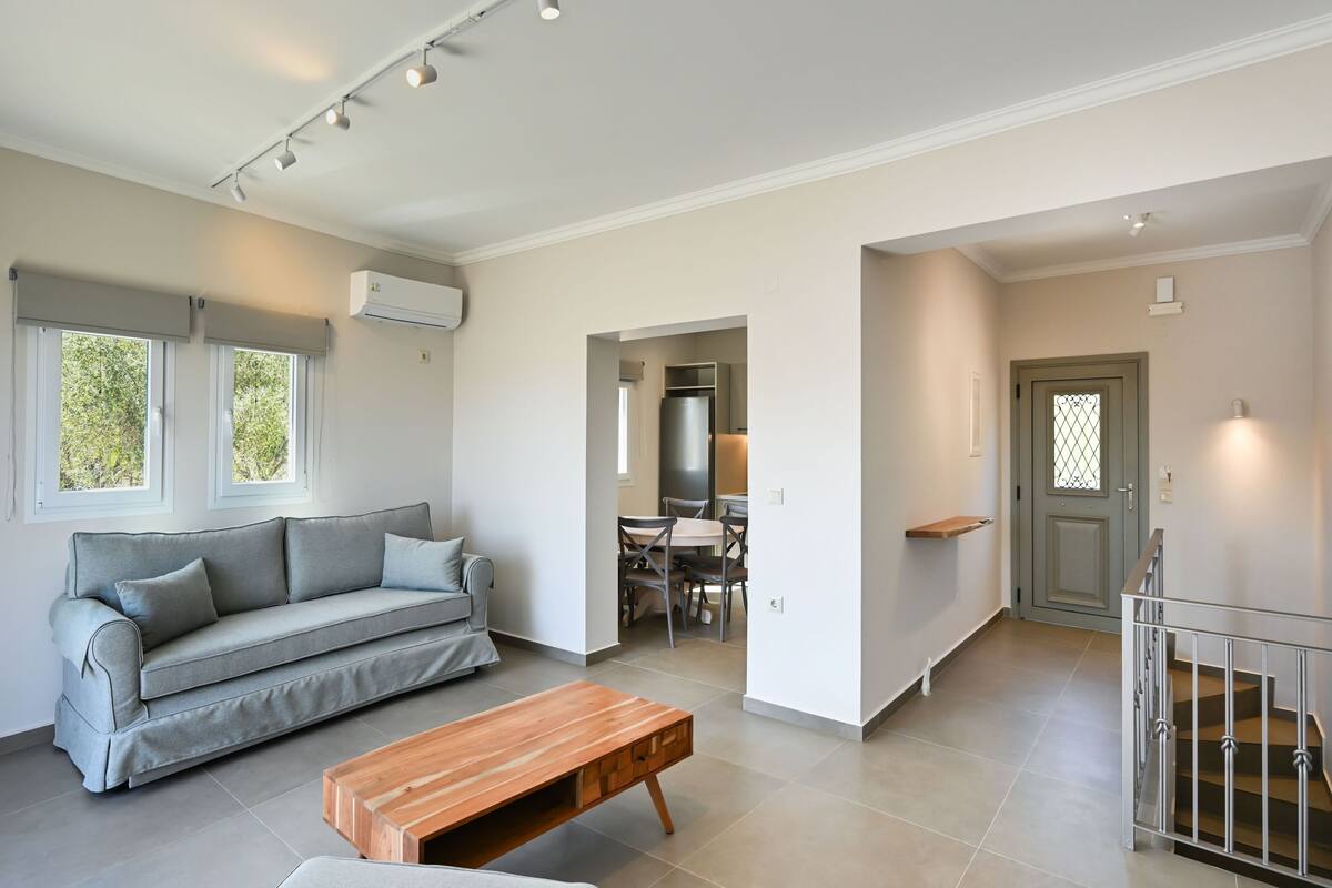 Un salon moderne avec un canapé gris, une table basse en bois et un coin repas comprenant une table et des chaises. Un escalier avec une rampe en métal mène à une porte.