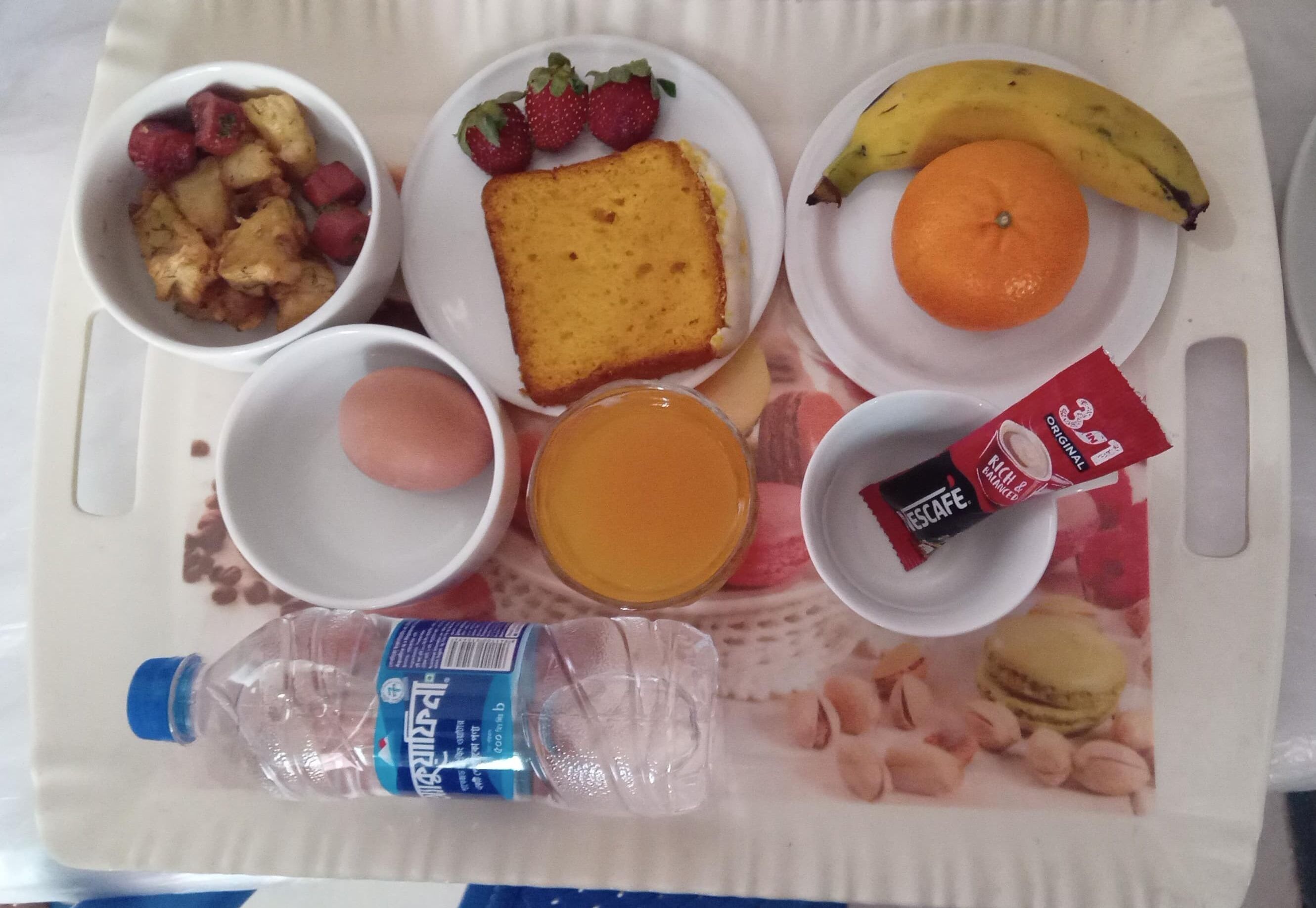Ein Tablett mit Speisen, darunter eine Wasserflasche, ein Glas Orangensaft, eine Scheibe Toast, Erdbeeren, eine Orange, eine Banane und eine Packung Instantkaffee.